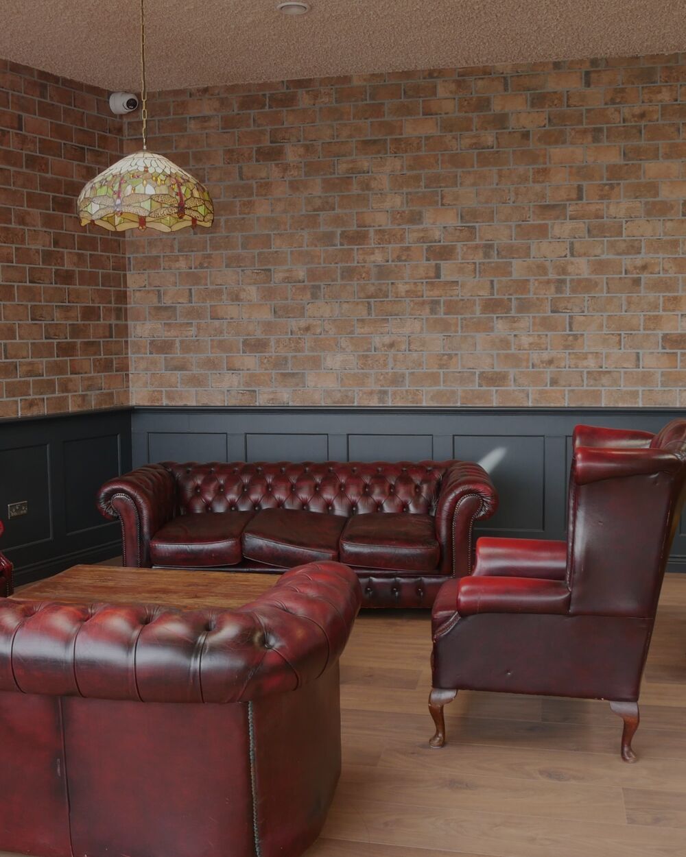 Brick-walled lounge with dark panelling, red leather Chesterfield sofas and armchairs, and a stained-glass pendant lamp.